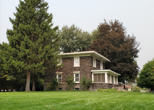 A two-story stone house surrounded by green grass and trees on a clear day.