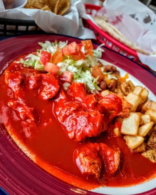 A plate of chicken in red sauce, served with lettuce, tomatoes, beans, and crispy potatoes.