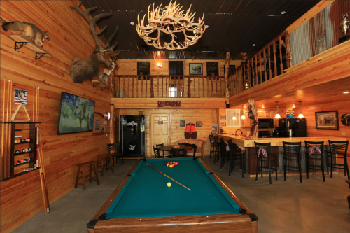 A rustic game room featuring a pool table, bar area, mounted animal heads, and wooden walls.