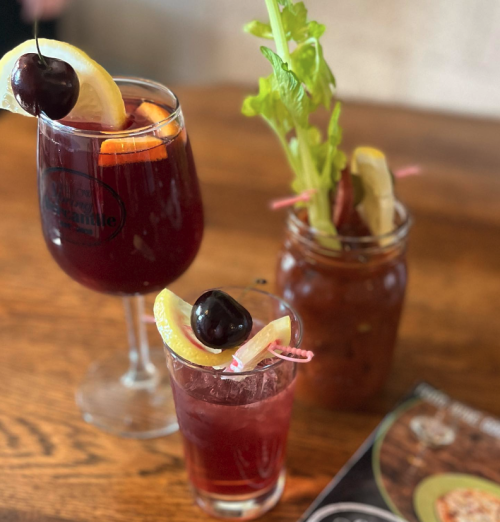 Three colorful cocktails on a wooden table: a large glass with fruit, a small glass with a cherry, and a jar with garnishes.