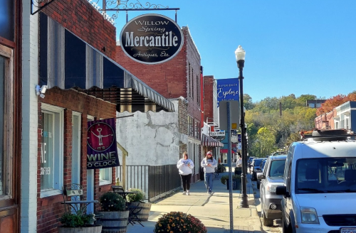 Two people walk along a charming street lined with shops and colorful signs on a sunny day.
