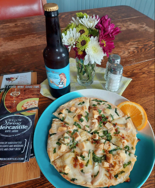 A plate of flatbread topped with chicken and herbs, a bottle of beer, and a small vase of flowers on a wooden table.