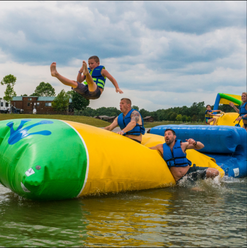 A child jumps off a large inflatable water slide into a lake, while others play on the slide nearby.