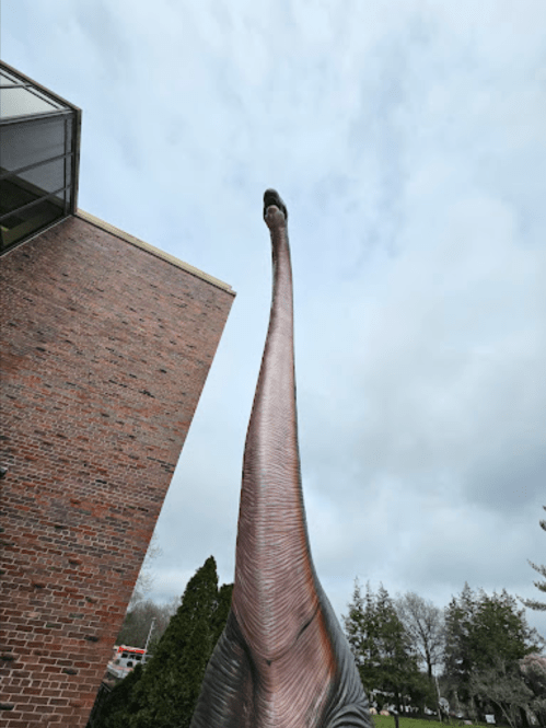 A tall dinosaur statue with a long neck, positioned next to a brick building under a cloudy sky.