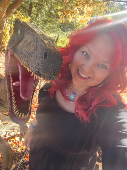 A woman with red hair smiles next to a dinosaur statue in a colorful autumn setting.