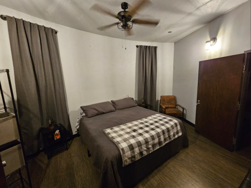 Cozy bedroom with a bed, gray bedding, a chair, and curtains, featuring a ceiling fan and warm lighting.