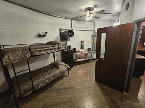 A small room with a bunk bed, a ceiling fan, and a door leading to another area. Minimal furnishings and decor.