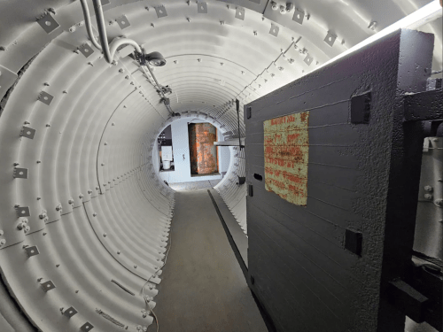 A long, cylindrical corridor with metal walls and a heavy door at the entrance, leading to a dimly lit space.