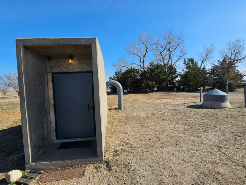 A concrete structure with a door stands in an open area, surrounded by trees and a circular tank in the background.