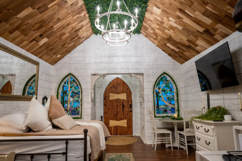 Cozy bedroom with wooden ceiling, stained glass windows, and rustic decor, featuring a bed, table, and decorative plants.