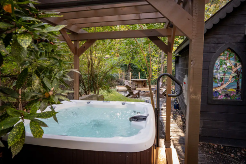 A serene outdoor hot tub surrounded by greenery and a wooden structure, with a glimpse of a garden and a cabin nearby.