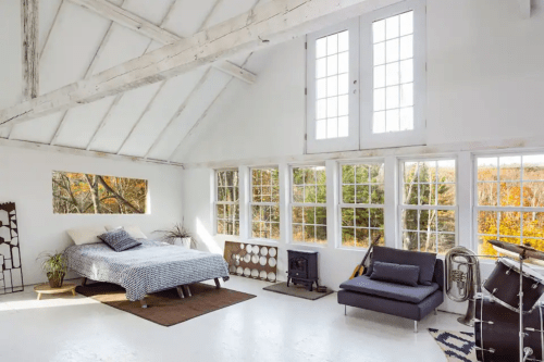 Bright, airy bedroom with large windows, a bed, a sofa, and musical instruments, surrounded by autumn foliage.