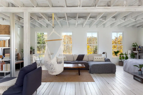 Bright, airy living room with white beams, a hammock, cozy seating, and large windows showcasing autumn foliage outside.