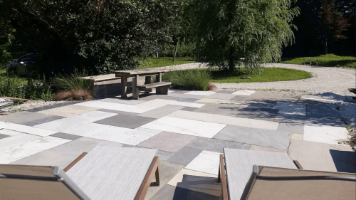A patio area with stone tiles, two lounge chairs, a wooden table, and greenery in the background.