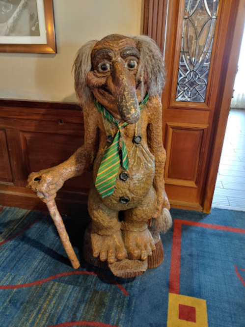 A whimsical wooden troll statue wearing a green striped tie, standing with a cane in a hotel lobby.