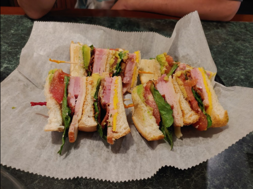 A platter of stacked sandwiches with layers of ham, cheese, lettuce, tomato, and avocado on white bread.