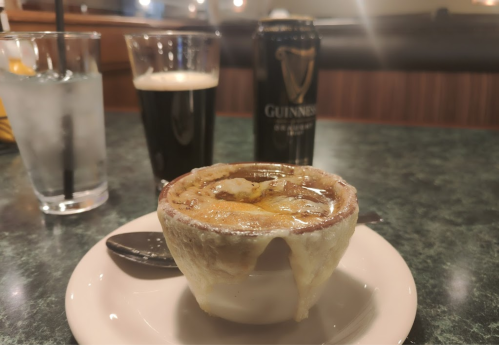 A bowl of French onion soup with melted cheese, a spoon, a glass of water, and a can of Guinness beer on a table.