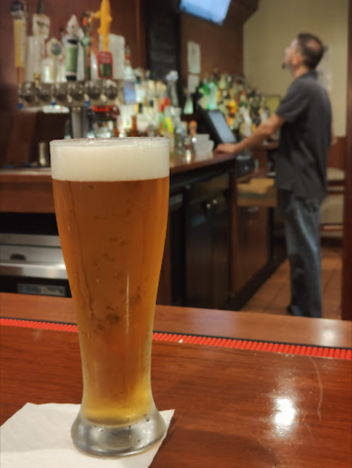 A glass of beer sits on a bar counter, with a blurred figure standing in the background near the bar.