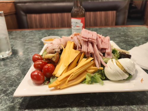 A plate of salad with sliced ham, turkey, cheddar cheese, cherry tomatoes, and a hard-boiled egg, with dressing on the side.