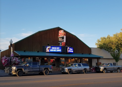 A rustic restaurant building with a green roof, featuring a sign and parked trucks in front. Bright blue sky above.