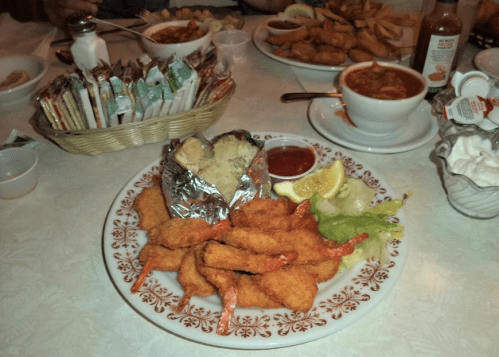 A plate of fried shrimp with a baked potato, lettuce, lemon, and dipping sauce, surrounded by various side dishes.