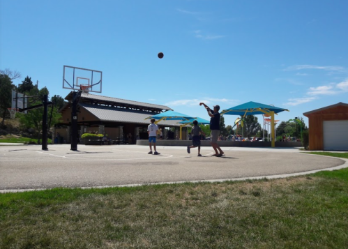 Three people playing basketball on an outdoor court under a clear blue sky with shaded areas in the background.