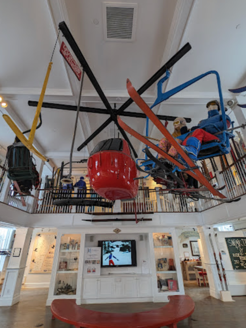 A whimsical display featuring ski lifts, skis, and mannequins in a museum setting, with a TV screen in the background.
