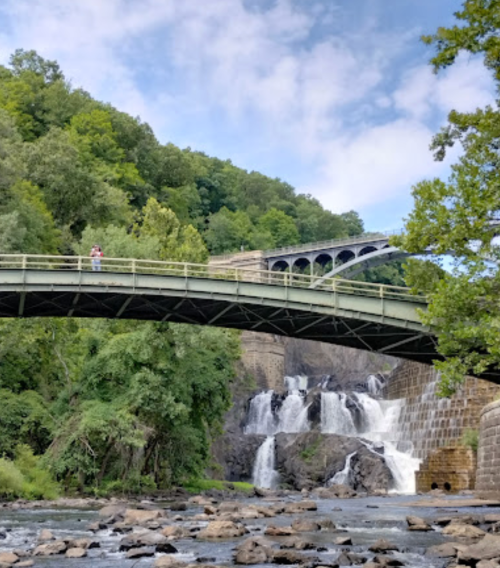 A scenic view of a bridge over a waterfall, surrounded by lush greenery and a clear blue sky.