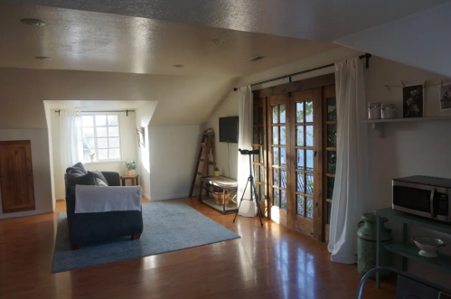 Cozy, well-lit living space with a sofa, wooden doors, and a small kitchenette area. Natural light from windows.
