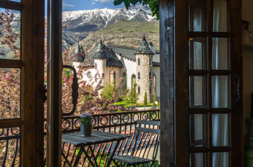 View from a balcony featuring a castle-like building and snow-capped mountains in the background, surrounded by blooming flowers.
