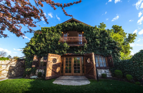 A charming house covered in greenery, featuring wooden double doors and a balcony under a blue sky.