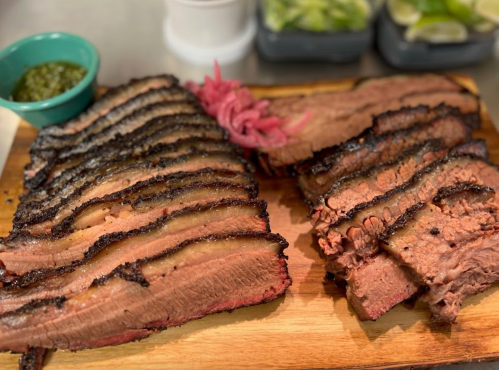 Sliced smoked brisket on a wooden board, accompanied by pickled onions and a small bowl of green sauce.