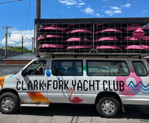A colorful van with "Clark Fork Yacht Club" painted on the side, topped with pink inflatable kayaks.
