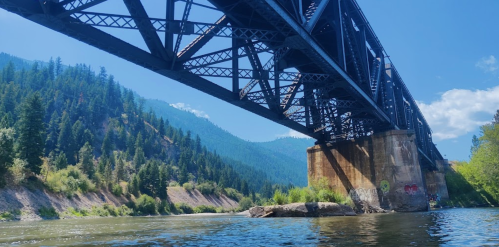 A steel bridge spans a river, surrounded by lush green hills and a clear blue sky.