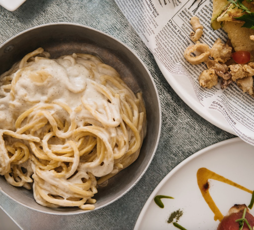 A bowl of creamy pasta sits on a table alongside a plate of fried seafood and a colorful dish.