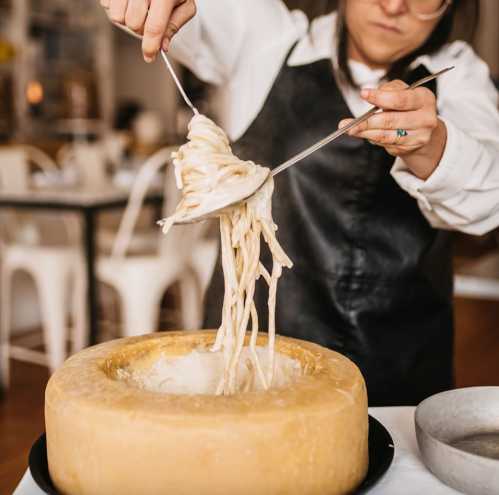 A person in an apron lifts spaghetti from a large wheel of cheese, steam rising in a dining setting.