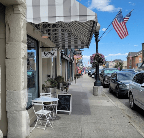A charming street scene with a café, American flags, and parked cars under a blue sky with fluffy clouds.