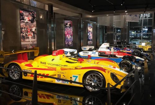 A display of colorful race cars in a museum, featuring a yellow Porsche and other racing vehicles in the background.
