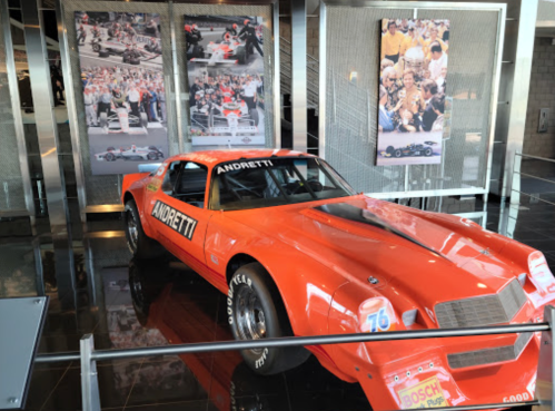 An orange race car with "ANDRETTI" on it, displayed in a museum alongside racing photos.