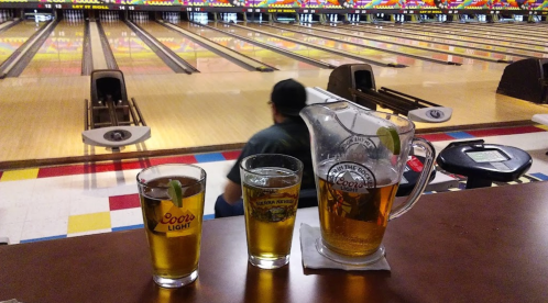 Three glasses of beer and a pitcher sit on a table, with bowling lanes visible in the background.
