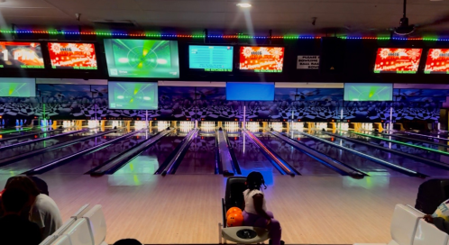 A bowling alley with multiple lanes, colorful lights, and screens displaying scores. A person sits with a bowling ball.