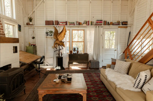 Cozy living room with a sofa, wooden furniture, bookshelves, and large windows, featuring rustic decor and warm lighting.
