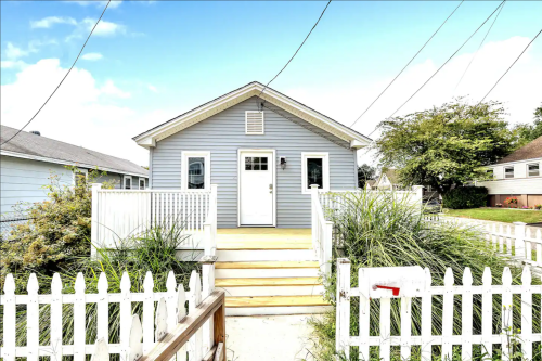 A light blue house with a white picket fence and steps leading to the front door, surrounded by greenery.