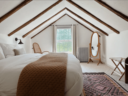 Cozy attic bedroom with a sloped ceiling, large window, wooden accents, and a warm-toned blanket on the bed.