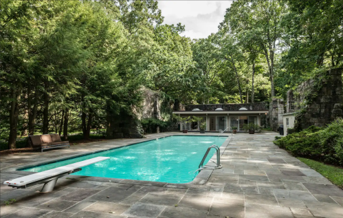 A serene outdoor pool surrounded by lush greenery and stone walls, with a diving board and lounge chairs nearby.