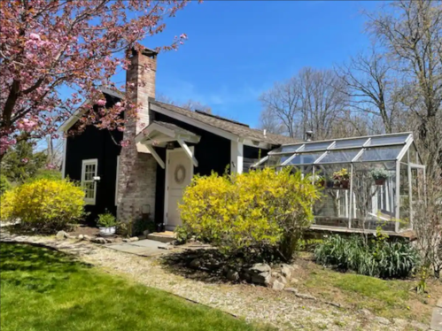 A charming black house with a stone chimney, surrounded by blooming trees and a greenhouse in a sunny garden.