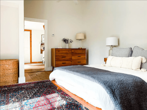 A cozy bedroom with a bed, decorative pillows, a wooden dresser, and a woven basket, featuring warm, neutral tones.