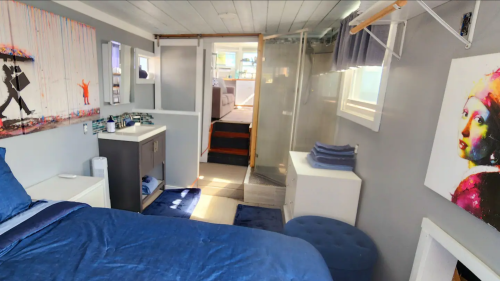 Cozy bedroom with blue decor, modern art, and a bathroom area, featuring a bed, storage, and natural light.