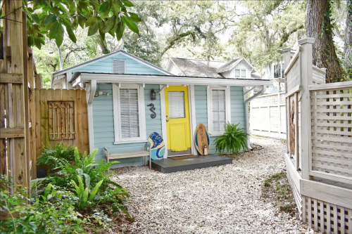 A charming blue cottage with a yellow door, surrounded by greenery and gravel pathways. Surfboards lean against the wall.