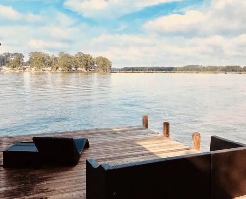 A serene lakeside view from a wooden dock, featuring calm water and distant trees under a partly cloudy sky.